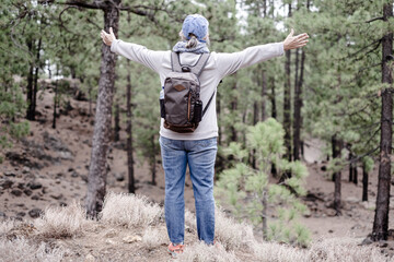 Back view of mature woman holding backpack standing in the pine forest with open arms enjoying nature and adventure. Sport and healthy lifestyle concept