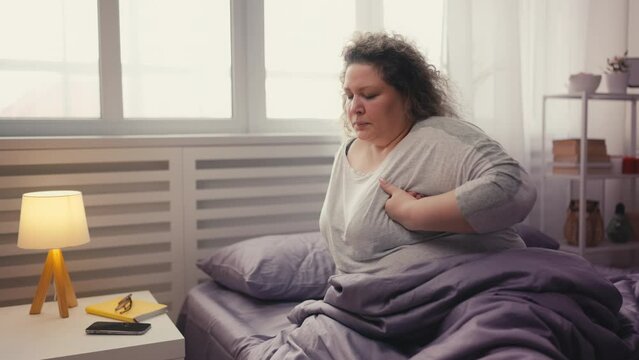 Overweight woman holding chest, sitting on bed and suffering pain, heart attack