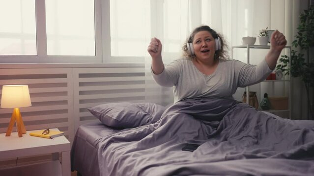 Cheerful overweight woman listening favorite song on mobile app, dancing in bed