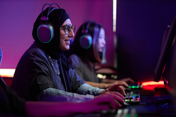 Side view portrait of Muslim young woman playing video games in cybersports club and smiling...