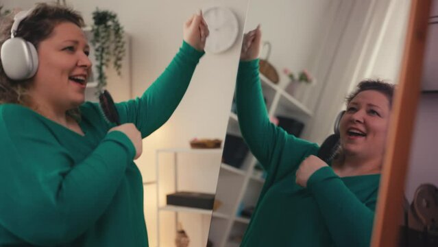 Funny body positive woman dancing in front of mirror, singing with brush in hand