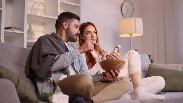 Boring young woman hold smartphone scrolling social media and ignoring her boyfriend or husband with popcorn laughing watching comedy movie show serial enjoying leisure time at home together