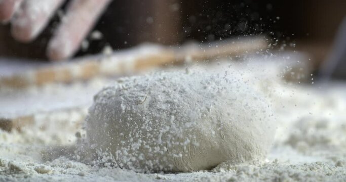Super slow motion close up of professional artisan baker chef sprinkles flour on raw loaf of dough while making homemade bread, pasta or pizza on rustic wooden table in traditional bakery kitchen.