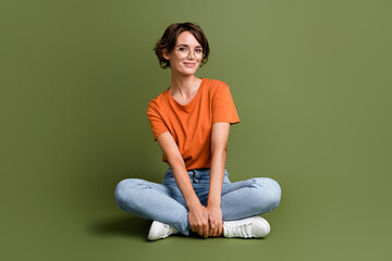Full size photo of friendly clever cute woman wear orange t-shirt jeans in glasses sitting on floor isolated on khaki color background