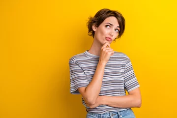 Poster Portrait of suspicious girl with short hairdo wear grey t-shirt hold finger on chin look empty space isolated on yellow color background © deagreez