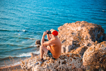 man sits on the top of a mountain and looks through binoculars at the seascape. Travel and tourism