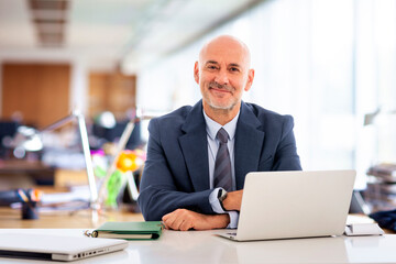 Executive businessman with his laptop sitting at office desk and looking at camera