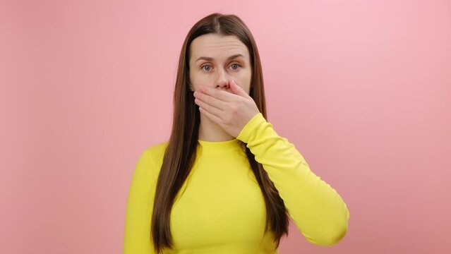 Portrait of afraid shocked young caucasian woman 25s wearing yellow sweater covering mouth with palm, keep secret, does not tell anyone, posing isolated over plain pink color background wall in studio