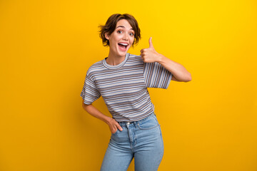 Portrait of overjoyed lovely funky girl wear grey t-shirt showing thumb up holding hand in pocket isolated on yellow color background