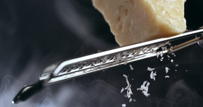 Super slow motion macro of a professional chef grating traditional parmesan cheese with steel grater on the dish close up and pieces of cheese fall at 1000 fps for preparing pasta.