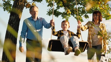 Caring grandma and grandpa push smiling grandson on swing enjoying time together