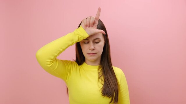 Portrait of upset disappointed young caucasian woman showing looser gesture with hand on forehead, wearing yellow sweater, posing isolated over pink color background wall in studio. Failure concept