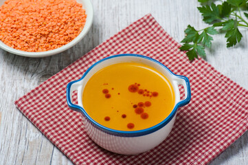 Top view of a bowl of classical Turkish red lentil soup with another bowl of uncooked red lentils and a twig of fresh parsley in the background on a white vintage wooden table