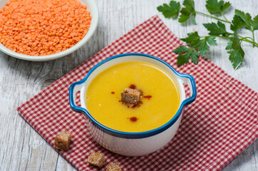 Top view of a bowl of classical Turkish red lentil soup with croutons, another bowl of uncooked red lentils and a twig of fresh parsley in the background on a white vintage wooden table