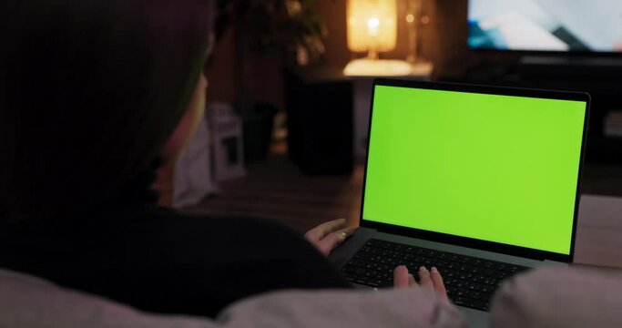 Freelance concept. Young woman with long hair sitting on couch in the living room watching tv typing working online holding laptop with green screen mock-up.