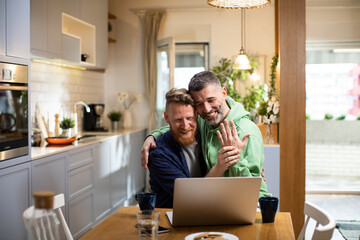 Happy gay couple showing engagement ring to video call at home