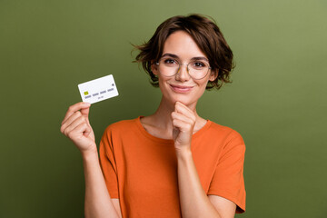 Photo of clever positive girl dressed orange t-shirt in glasses holding credit card arm on chin isolated on khaki color background - obrazy, fototapety, plakaty
