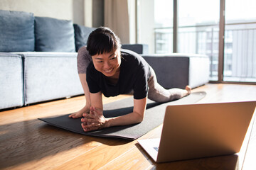 Smiling Asian woman stretching doing yoga online class from laptop at home in living room
