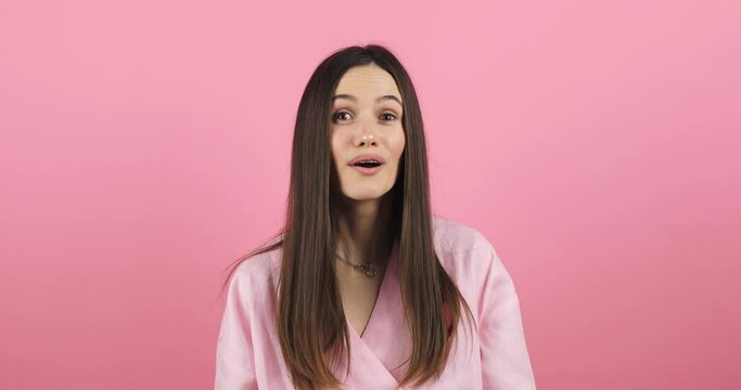 Attractive young woman look by side, turn her head by side to camera and seen somebody, she look happy, smile, waving her hand wear pink dress isolated on pink background. Happy to see you.