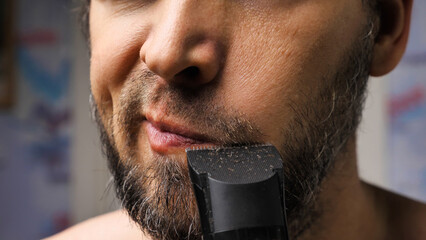 A man trims his beard with a trimmer. Close up. An electric razor trimming a beard. Black beard with grey hairs. Beard care at home. - obrazy, fototapety, plakaty