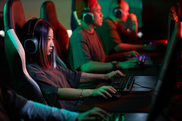 Obraz premium Side view portrait of young Asian woman playing video games in cybersports club and wearing pro headphones, copy space