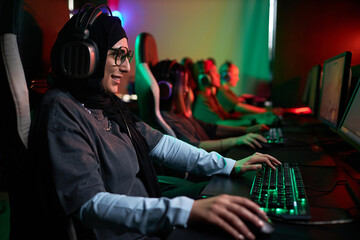 Fototapeta na wymiar Side view portrait of smiling Muslim woman playing video games in cybersports club with neon lighting, copy space