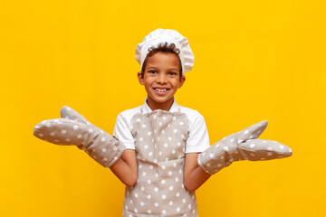 boy african american baker in uniform and chef's hat holding empty hands on yellow isolated background