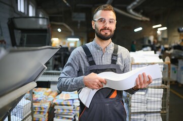 Printing house interior with modern offset print machine and operator in working uniform checking...