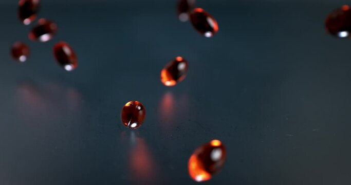 Slow motion macro of transparent red soft gel vitamins and nutritional supplements capsules falling on grey mirroring surface in scientific pharmaceutical laboratory.