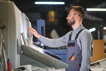 Printing house interior with modern offset print machine and operator in working uniform checking...