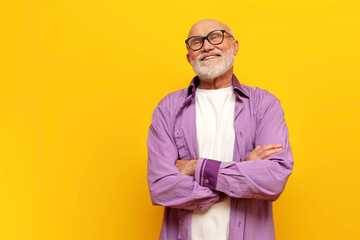 pensive old bald grandfather with gray beard in eyeglasses stands with his arms crossed on yellow...