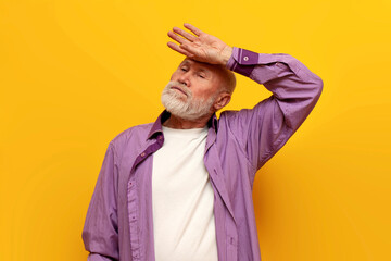 tired old bald grandfather in a purple shirt wipes sweat from his forehead on yellow isolated...