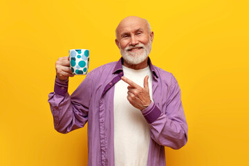 old bald grandfather in purple shirt holds cup of drink and points on yellow isolated background