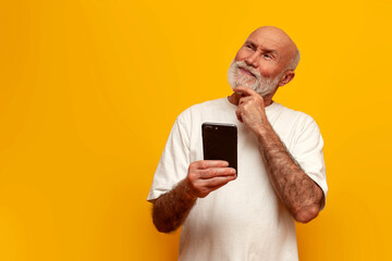 pensive old bald grandfather in white t-shirt uses smartphone and thinks on yellow isolated...