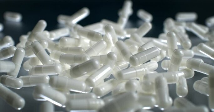 Slow motion macro of white painkiller or antibiotic capsules falling on grey mirroring surface in scientific pharmaceutical laboratory.
