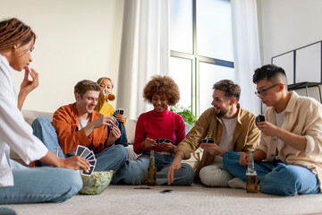 multiracial group of young people sitting at home with beer and popcorn and playing cards with...