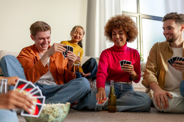 multiracial group of young people sitting at home with beer and popcorn and playing cards with...