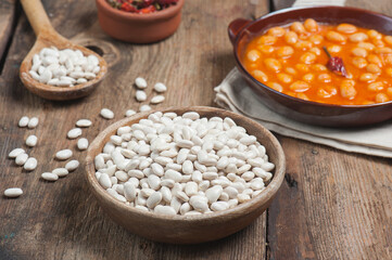 High angle photo of a bowl of navy beans on a vintage wooden table with traditional home cooked navy beans dish on a linen napkin in the background