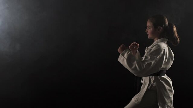 Young karate girl wearing kimono and posing against black background with smoke. Slow motion recorded at 100fps