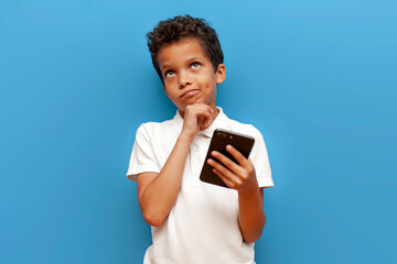 pensive african american boy in white polo using smartphone dreams and imagines on blue isolated background - obrazy, fototapety, plakaty
