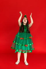 A girl in a festive carnival costume of little Christmas tree pointing at an empty space on a red background. Winter holidays concept. Sale and advertising