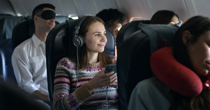 Beautiful blonde girl travels by plane and listens to music while watching phone, relaxing happy and carefree during the flight in first class with hi-speed internet technology. 