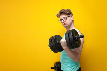 non-athletic guy in glasses lifts heavy dumbbells on a yellow background, a nerd trains and goes in...