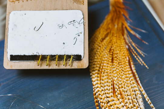 Brown feathers on a blue table used for tying fishing flies, next to a row of tied flies and hooks
