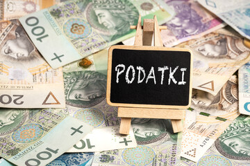 a small wooden writing board standing on scattered Polish zloty PLN banknotes, a chalk inscription "Podatki" on the black board, translation: taxes (selective focus)