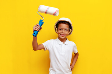 african american boy painter repairman in hard hat and safety glasses holds roller and paints on blue background