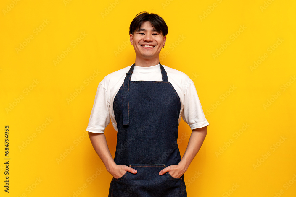 Wall mural asian young male waiter in apron standing and smiling on yellow isolated background, korean guy barista businessman