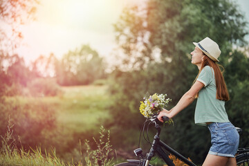 A young woman in a hat with a bouquet in her hand holds a bicycle in the park. A beautiful girl...