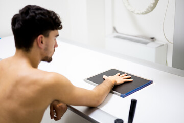 An unrecognizable young man sits in a hospital room undergoing an x-ray test on his hand to detect fractures, tumors, foreign objects or degenerative hand disorders.