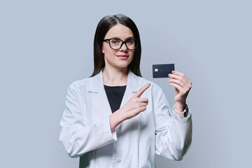 Young woman medical specialist, doctor holding bank credit card on gray background